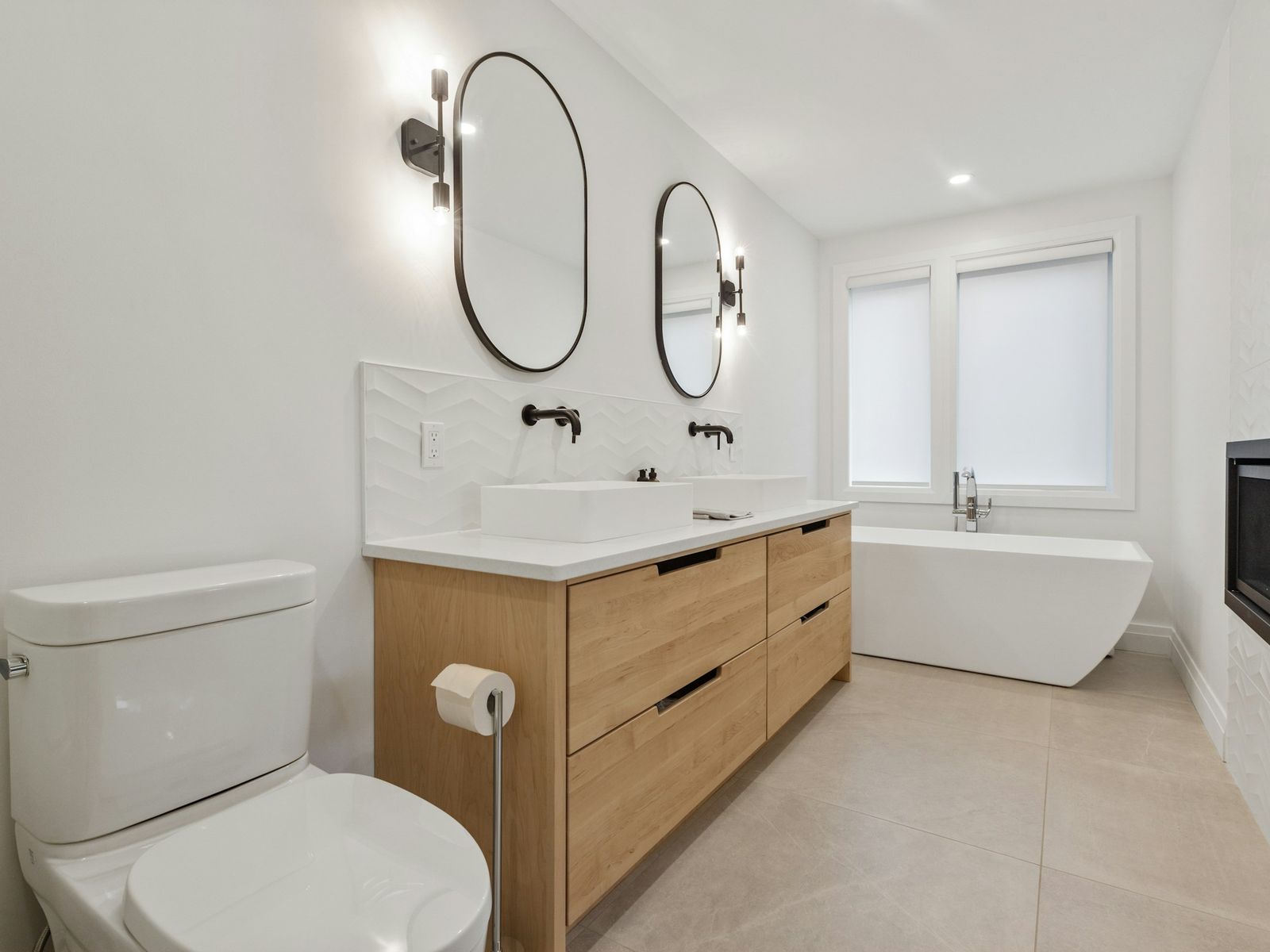 Light-wood double vanity with matte black wall faucets and a freestanding tub, illustrative stock photo